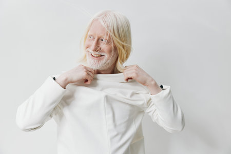 Stylish elderly man with long blonde hair smiling and adjusting his white shirt, set against a minimalist white background, exuding confidence and positivityの写真素材