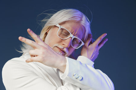Stylish elderly man with long white hair and glasses, striking a playful pose against a dark blue background, showcasing confidence and uniqueness in modern fashionの写真素材
