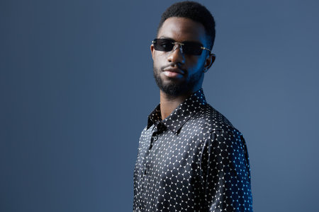 Stylish african man in polka dot shirt and sunglasses posing confidently against a vibrant blue backgroundの写真素材