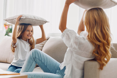 Mother and daughter having fun with pillows on their heads, sitting together on a cozy couch in a bright living roomの写真素材