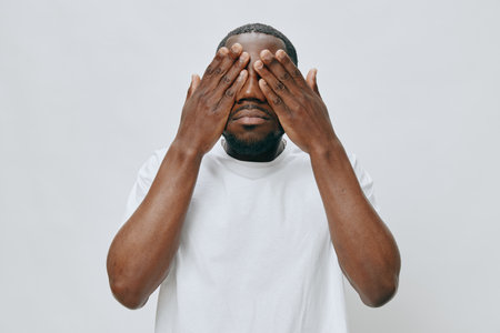 Emotional portrait of a man covering his eyes in a plain, neutral background, expressing introspection and contemplation while wearing a simple white t shirt, creating a powerful visual impactの写真素材