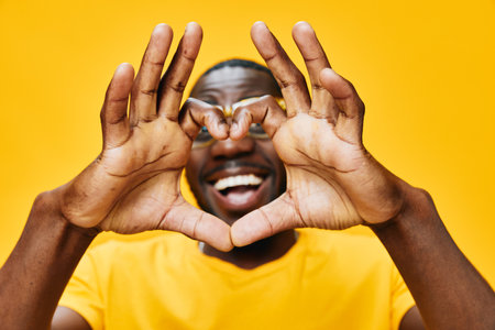 Happy man making heart shape with hands, wearing yellow shirt, smiling, vibrant yellow background conveying joy and positivity through playful expression and energetic vibeの写真素材