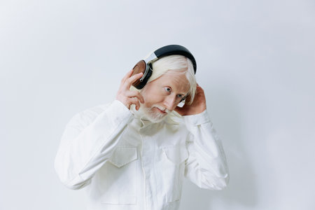 Elderly man enjoying music with headphones on a plain white background, wearing a stylish all white outfit that highlights his silver hair and beardの写真素材