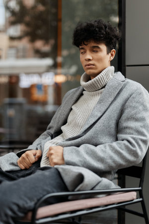 Fashionable young man in sweater and jeans sitting on a chair in front of a window with hands on kneesの写真素材