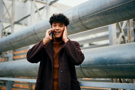 Fashion man in brown coat using cell phone in industrial setting with pipes in backgroundの写真素材