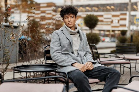 Fashionable young man in a grey coat sitting in front of a building with trees in the backgroundの写真素材