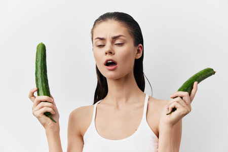 Woman holding cucumbers with a playful expression on a white background, emphasizing freshness and health The image captures a humorous and light hearted concept about vegetablesの写真素材