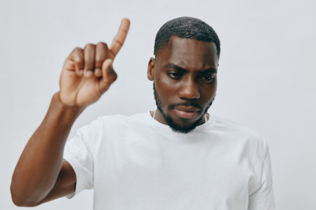 Young man expressing a thoughtful gesture with one finger raised, wearing a plain white t shirt, set against a neutral light background highlighting his focus and determinationの写真素材