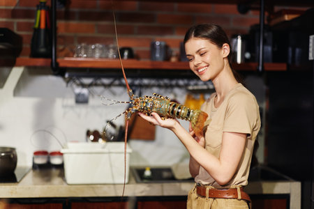 Young woman holding a lobster in a restaurant kitchen and smiling at the camera, focus on fresh seafood conceptの写真素材