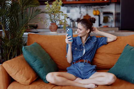 Young woman relaxing on orange couch with feet on coffee table, using cell phone in living roomの写真素材