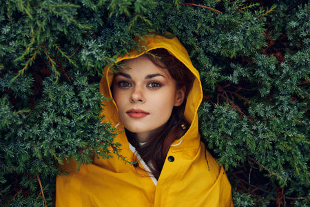 Mysterious young woman in a vibrant yellow raincoat hiding behind lush evergreen trees in a remote forestの写真素材