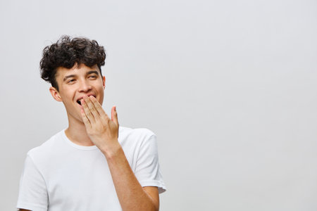 Smiling young man with curly hair in a white t shirt, covering his mouth with his hand, expressing joy or amusement against a light gray backgroundの写真素材