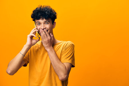 Young man talking on phone, surprised expression, bright orange background, casual style, happiness concept, communication, youth, cheerful mood, emotions, connecting with othersの写真素材