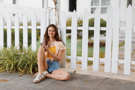 Young woman sitting by a white fence holding a coffee cup, wearing casual summer clothes and smiling while enjoying a sunny day in a pleasant outdoor settingの写真素材