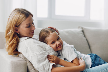 Mother and daughter bonding on a cozy living room couch in front of a sunlit window, sharing a warm...