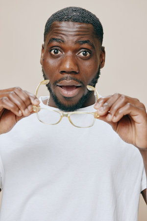 Surprised young man holding glasses, brown skin, casual white t shirt, expressive emotion, neutral...
