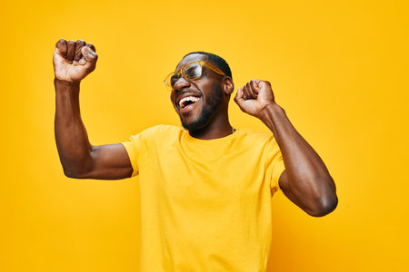 Happy man celebrating with joyful expression, wearing a yellow t shirt and sunglasses against a bright yellow background, embodying positivity and vibrant energyの写真素材