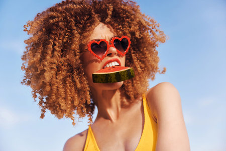 Woman with curly hair wearing heart shaped sunglasses holds a slice of watermelon against a blue sky backgroundの写真素材