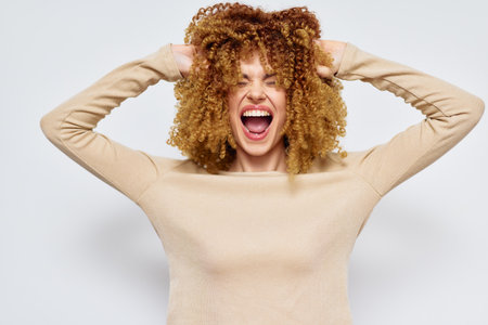 Joyful woman with curly hair expressing happiness in a light beige sweater against a soft pastel background, showing vibrant emotions and positivityの写真素材