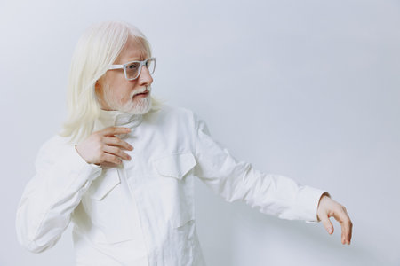 Elderly man with long white hair and glasses, dressed in a white jacket against a light gray background. He is gesturing thoughtfully, exuding a calm and reflective mood.の写真素材