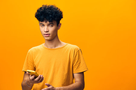 Young man with curly hair holding a yellow phone, standing against a bright orange background The vibrant colors create a playful and youthful atmosphere, emphasizing modern technologyの写真素材