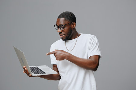 Young man using a laptop, wearing glasses, casual white t shirt, displaying excitement, neutral gray background. Modern technology, lifestyle, and engagement with digital devicesの写真素材