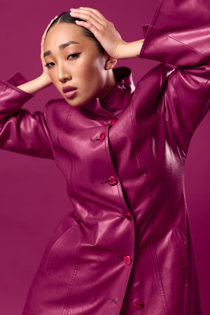 Fashionable asian woman in purple leather jacket posing with hands on head against matching background, hair pulled backの写真素材