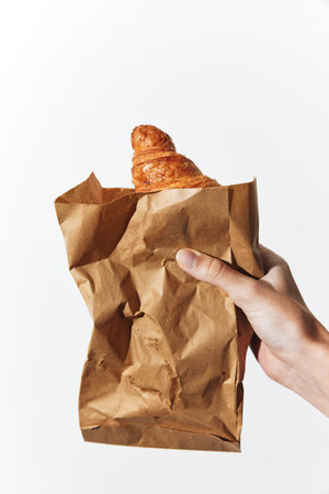 Person holding a brown paper bag with a croissant on a white backgroundの写真素材