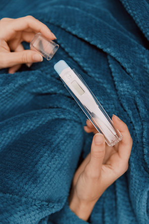Sick woman lying on blue bedding holding a thermometerの写真素材