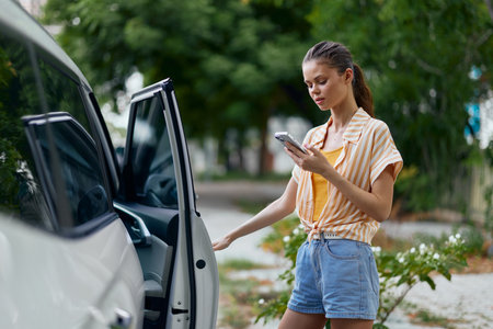 woman using smartphone by car, outdoor setting, casual outfit, warm weather, summer vibe, urban environment, greenery background, stylish appearance, responsible use of technologyの写真素材