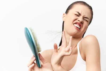 Woman experiencing frustration while brushing hair, looking annoyed with tangled strands Bright white background enhances the focus on her expression and the hairbrushの写真素材