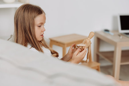 Young girl on couch playing with wooden doll while watching something on a laptop, cozy home settingの写真素材