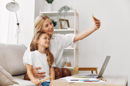 Mother and daughter collaborating on a laptop and paper while sitting on a cozy couch in a modern living roomの写真素材
