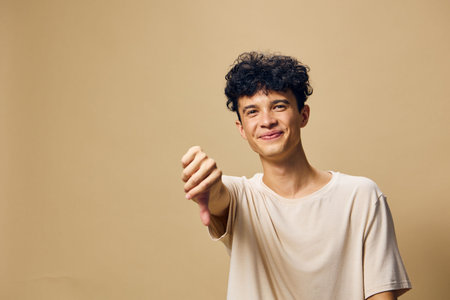 Young man expressing a thumbs down gesture in a neutral pose against a soft beige background, conveying disappointment or disapproval in a casual settingの写真素材