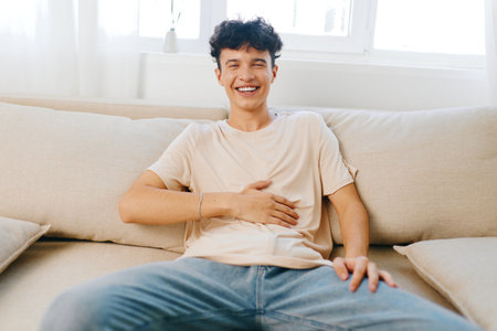 Young man laughing and resting on a light colored sofa indoors, showcasing a joyful expression and casual attire Bright and airy home environment with natural lightの写真素材