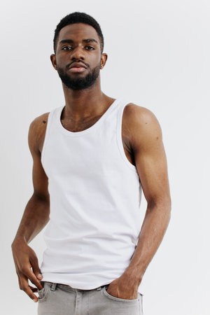 Confident young black man in casual white tank top and jeans standing with hands on hips against a neutral backgroundの写真素材