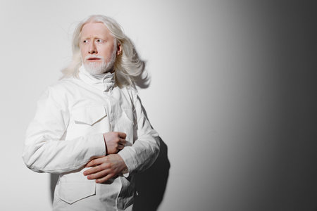 Elderly man with long white hair in a stylish white outfit standing against a gradient gray background, expressing a sense of contemplation and introspection with a soft poseの写真素材