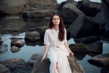 Serene woman in white dress sitting on ocean rock with rocky backgroundの写真素材
