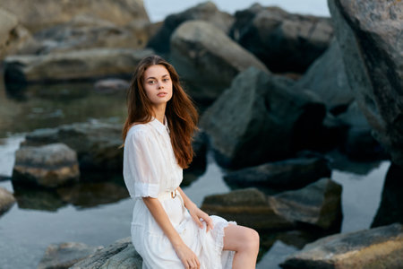 Serene woman in white dress sitting on rocky shore, gazing out at tranquil waterの写真素材