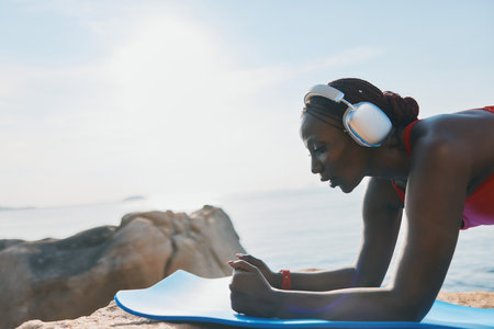 Woman doing plank exercise on a yoga mat by the ocean at sunrise, wearing headphonesの写真素材