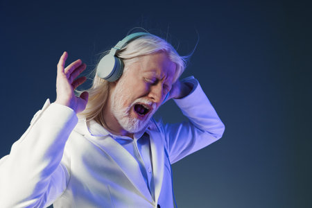 Elderly man enjoying music, wearing headphones, with a joyful expression against a dark blue background, showcasing a modern lifestyle and the joy of musicの写真素材