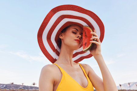 Woman in a yellow swimsuit holding a watermelon slice, wearing a large red and white striped hat, against a clear blue skyの写真素材