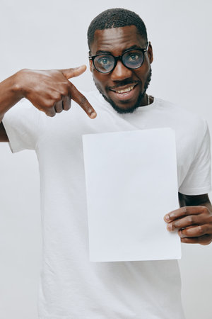 smiling man with blank sheet of paper, casual attire, pointing gesture, white background, cheerful expression, vibrant energy, studio lighting, self promotion, African descentの写真素材