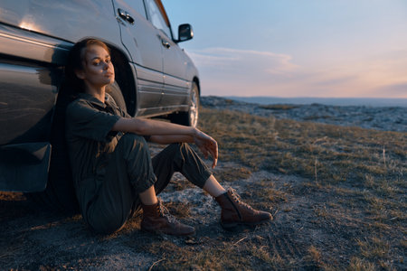 Woman sitting on car in the middle of field at sunset, contemplating the beauty of nature and seeking peace and tranquilityの写真素材
