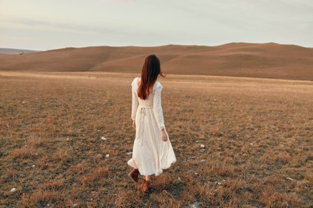 Woman in white dress walking through open field at sunset in travel and beauty conceptの写真素材