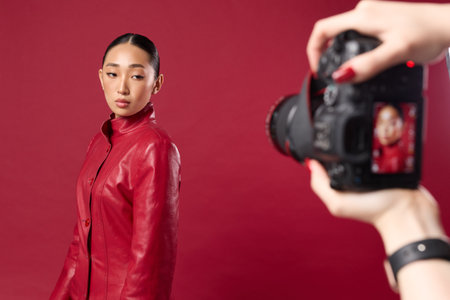 Fashionable asian brunette woman in red leather jacket taking a photo with digital camera on red backgroundの写真素材