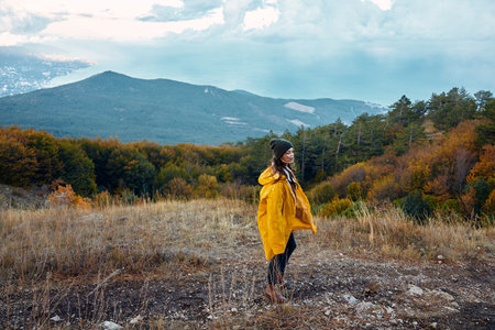 Woman in a vibrant yellow raincoat standing on a hill admiring the picturesque valley and majestic mountains landscapeの写真素材
