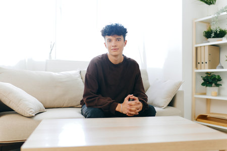 Young man sitting on a couch in a bright, minimalistic room, showcasing a relaxed atmosphere and modern decor around him with soft colors, ideal for lifestyle themesの写真素材