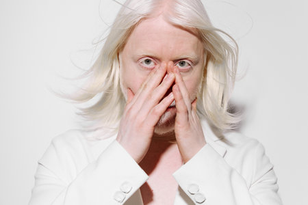 Portrait of a person with light blonde hair and pale skin, covering their face with hands, against a minimalistic white background, conveying emotions of vulnerability and introspectionの写真素材
