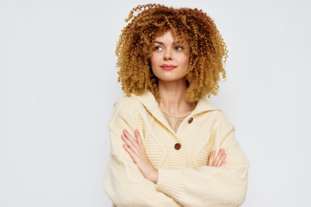 Confident young woman with curly hair wearing a cozy light sweater, standing against a plain white background, exuding warmth and self assurance in a relaxed poseの写真素材